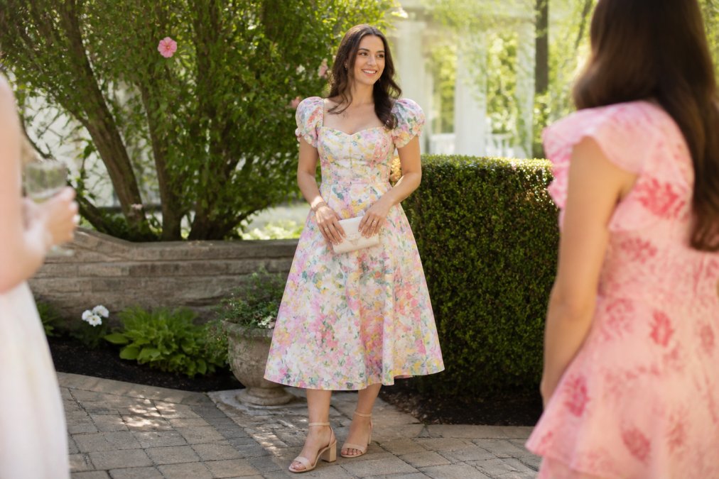 A woman in a pastel floral midi dress and block heels standing in a sunlit garden, holding a small clutch, smiling naturally