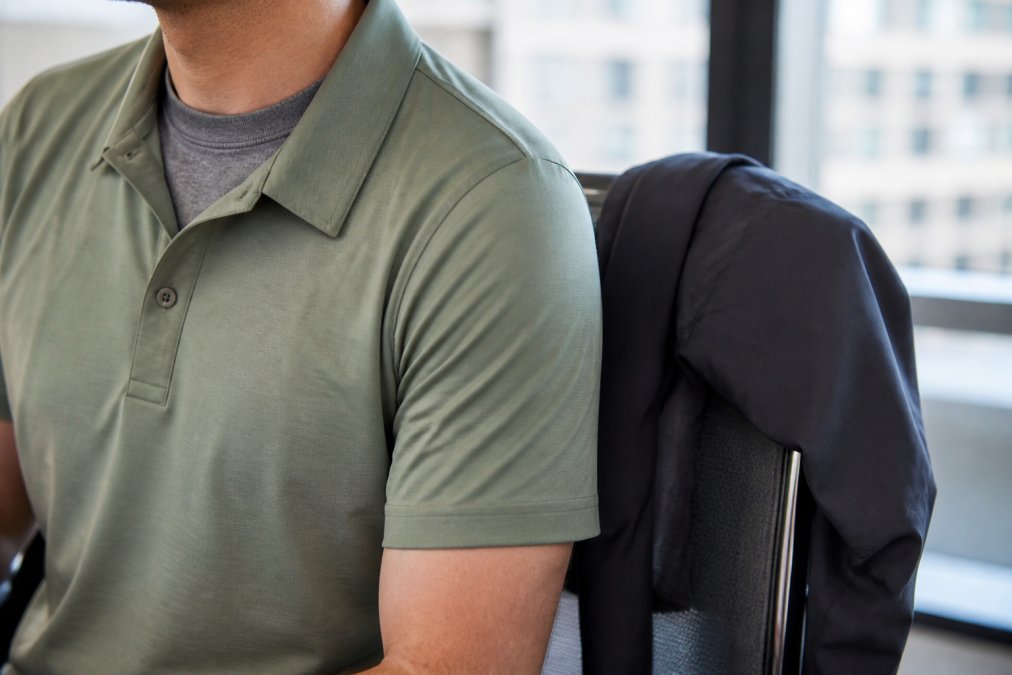Close-up of a man's torso showing layered athletic pieces: a moisture-wicking gray base layer visible at the neckline, a breathable sage-green performance polo shirt layered over it, and a lightweight charcoal performance jacket draped over the back of a modern office chair, with soft natural window light illuminating the fabric textures