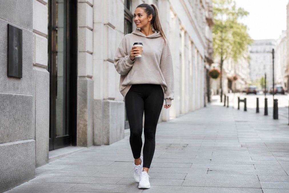 Woman wearing high-waisted black gym leggings styled with an oversized neutral-toned hoodie and white trainers, holding a coffee cup while walking through a bright urban street with modern buildings