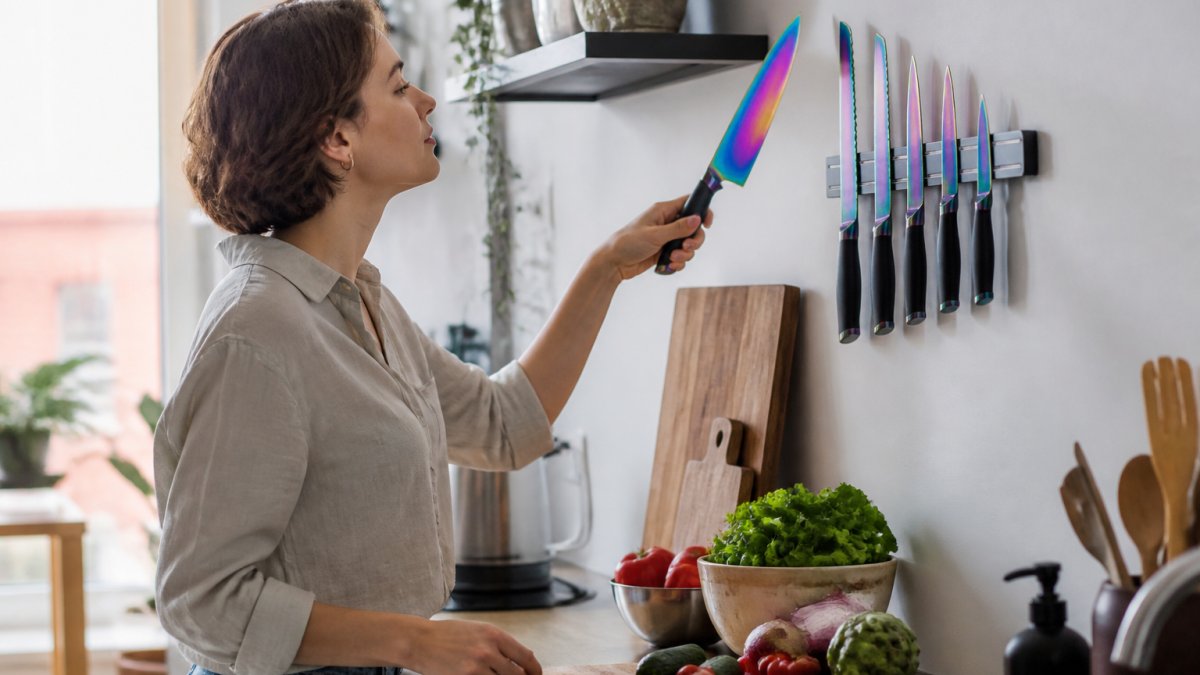 A young adult in a small bright apartment kitchen lifting an iridescent titanium-coated chef knife from a magnetic rack mounted on a white wall, with a wooden cutting board and fresh vegetables on the counter nearby