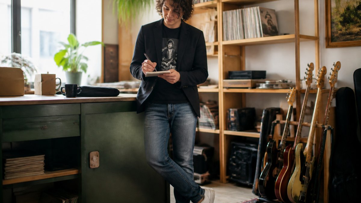 Person wearing a band tee layered under a structured blazer or denim jacket, styled with fitted jeans and clean sneakers, standing in modern casual setting with natural lighting