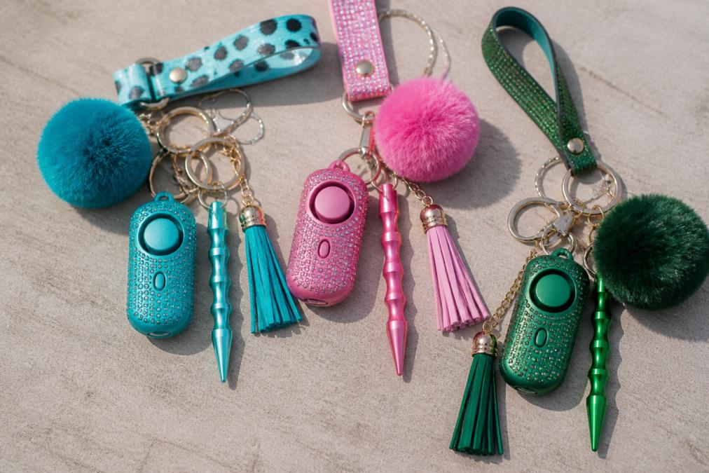 Close-up of colorful safety keychains arranged on a neutral surface, showing rhinestone details, pom-poms, wristlets, and various color palettes including turquoise cow print, pink rhinestones, and emerald green, with personal alarms and emergency tools visible