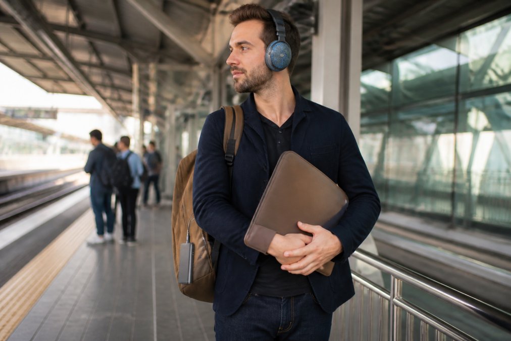 Professional commuter at a modern transit station holding a stylish laptop sleeve, wearing noise-canceling headphones with a transport-themed case, with a portable charger clipped to their bag, natural morning lighting