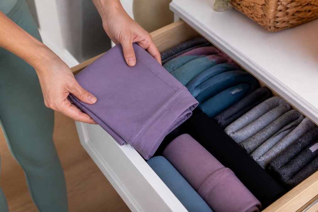 Hands carefully folding yoga leggings and placing them in a drawer with other neatly organised activewear, showing proper storage technique with soft lighting
