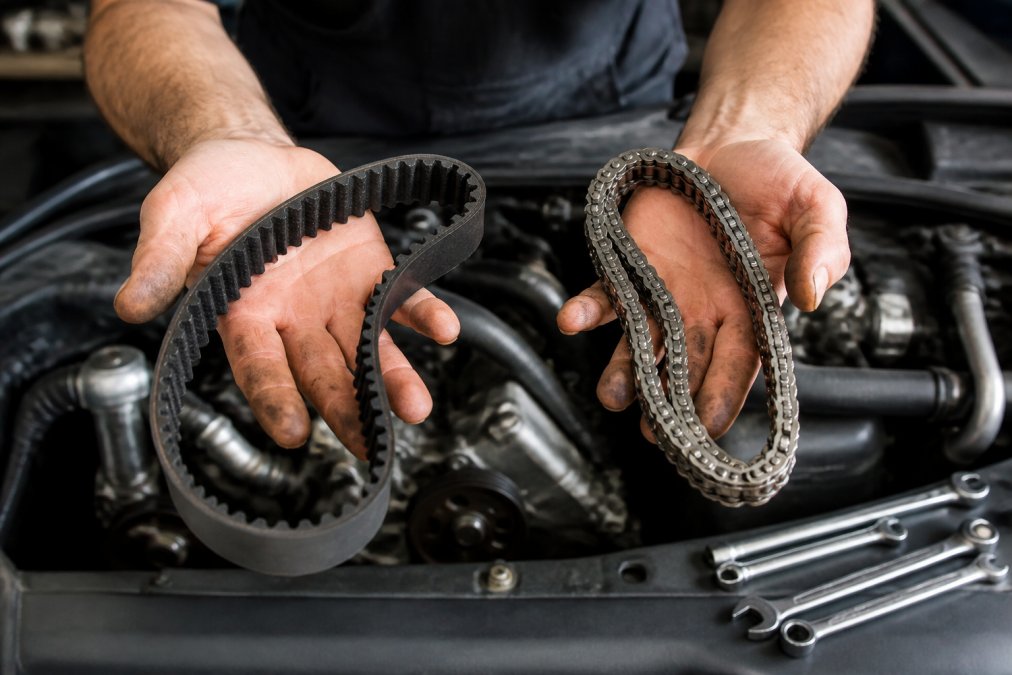 Professional mechanic's hands holding a rubber timing belt and metal timing chain side-by-side in an engine bay, with tools visible nearby and natural workshop lighting illuminating the contrast between the flexible belt and rigid chain components