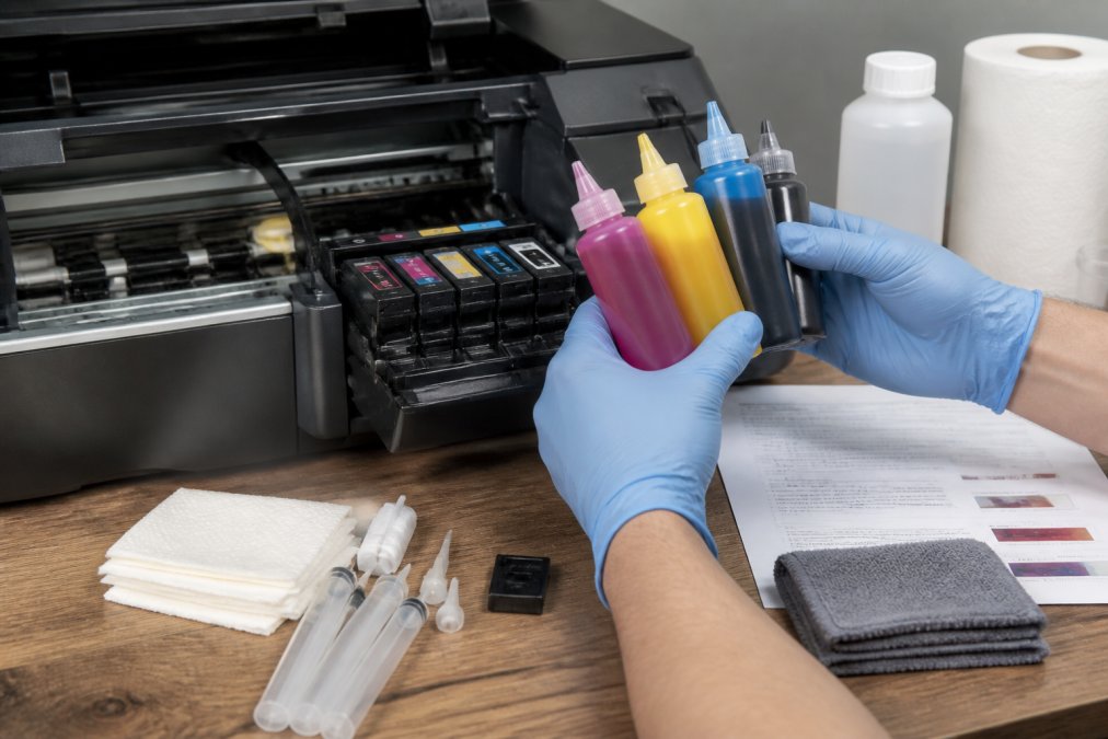Hands holding sublimation ink bottles over an open printer cartridge bay, with cleaning supplies, documentation, and lint-free cloths visible on a workbench