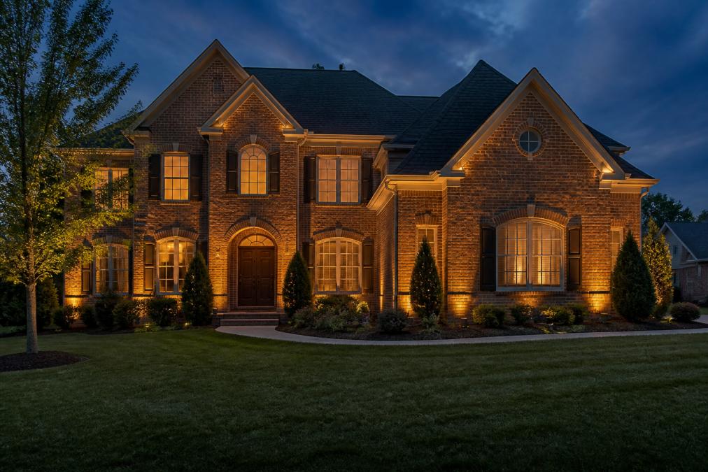 A two-story brick home at dusk with warm light grazing the facade from ground-level fixtures, highlighting the texture of each brick while the windows glow softly from within