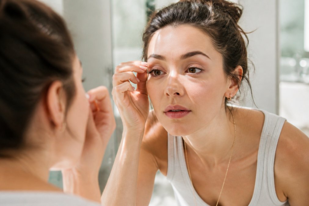 Close-up of a woman's face with naturally styled, subtle eyelash extensions, looking into a mirror in a bright, modern bathroom, soft morning light on her skin