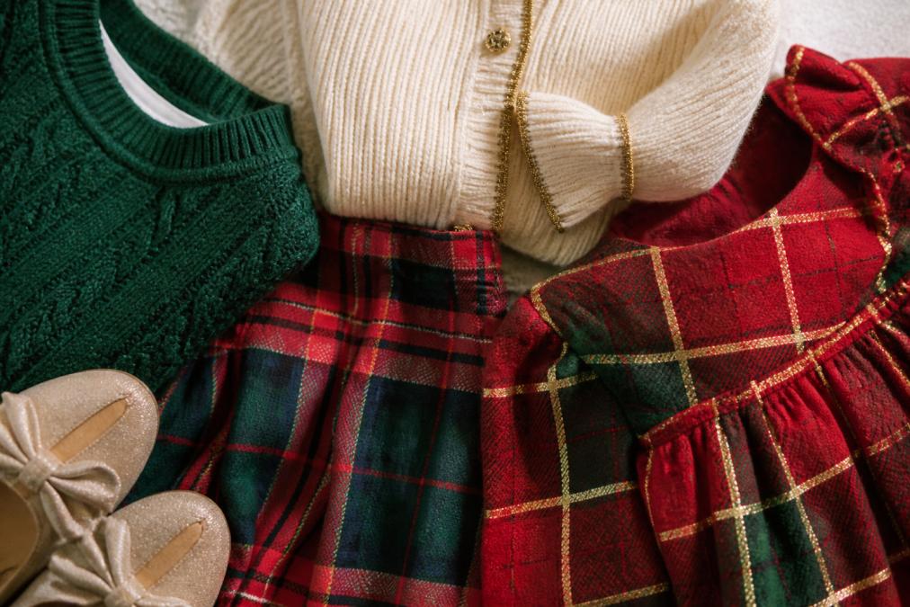 Close-up detail shot showing layered holiday outfits with textured plaid fabrics, cream ribbed trim, and subtle gold accents arranged on a neutral background
