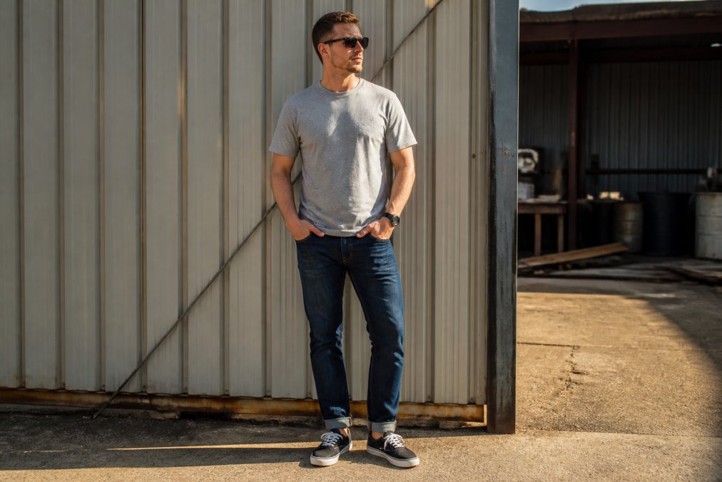 Man wearing a subtle motocross tee in neutral color with well-fitted jeans and casual canvas sneakers, standing relaxed outdoors in natural daylight, demonstrating how motocross apparel transitions to everyday wear