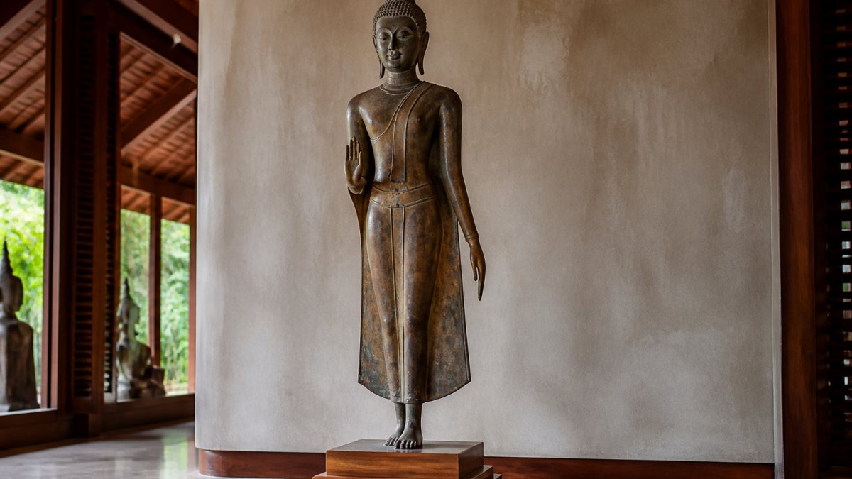 A serene Sukhothai-style walking Buddha statue with elongated limbs and smooth modeling, displayed on a wooden plinth in a softly lit gallery with warm natural light