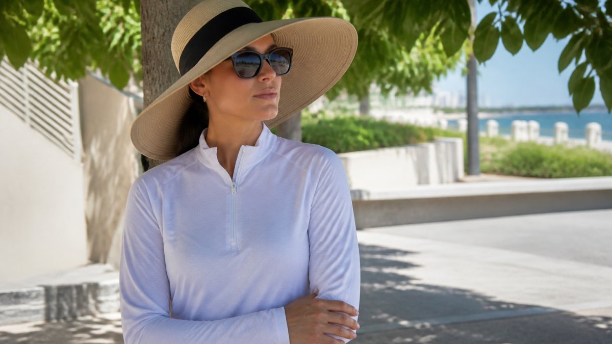 Person wearing a wide-brimmed hat with at least 3-inch brim, long-sleeved UPF-rated shirt in light fabric, UV-blocking sunglasses, and standing in shade during bright afternoon sunlight