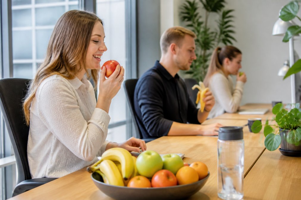 Medarbejdere på et moderne kontor, der spiser frisk frugt ved deres skriveborde, smilende og fokuserede, naturligt lys fra vinduerne, frugtskål på bordet