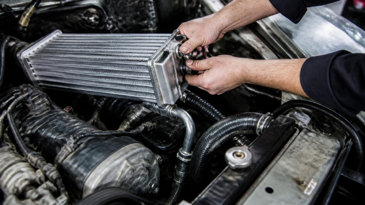Professional technician's hands installing a water-cooled supercharger heat exchanger on a truck engine, with coolant lines being connected to the manifold and radiator hoses visible, bright workshop lighting