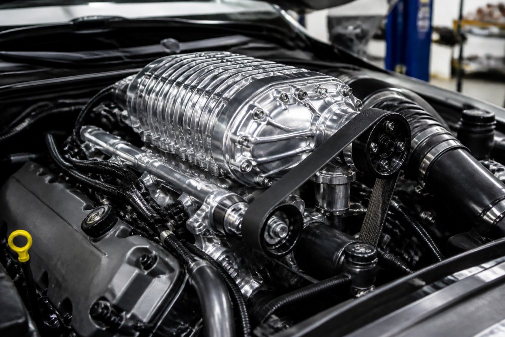 Close-up of a twin-screw supercharger unit mounted on a modern V8 engine in a clean workshop, showing the polished intake manifold and serpentine belt system under bright work lights