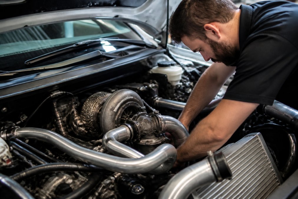 Technician working on turbocharger exhaust manifold installation with visible custom piping, intercooler connections, and wastegate assembly in an engine bay