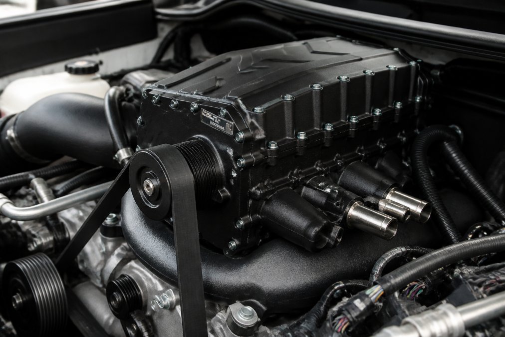 Close-up of a truck engine bay showing a twin-screw supercharger installed on the intake manifold with visible belt drive system and polished aluminum components