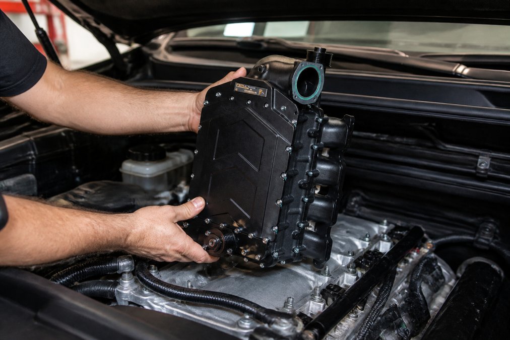 Experienced mechanic performing bolt-on supercharger installation on a truck engine, showing the straightforward fitment of the supercharger body to the intake manifold with visible belt and pulley assembly, no cutting or welding required