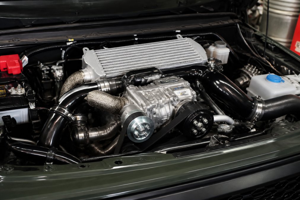 Close-up of an engine bay showing a supercharger mounted on top of an engine with visible belt, pulleys, and intercooler piping in a well-lit detail shot
