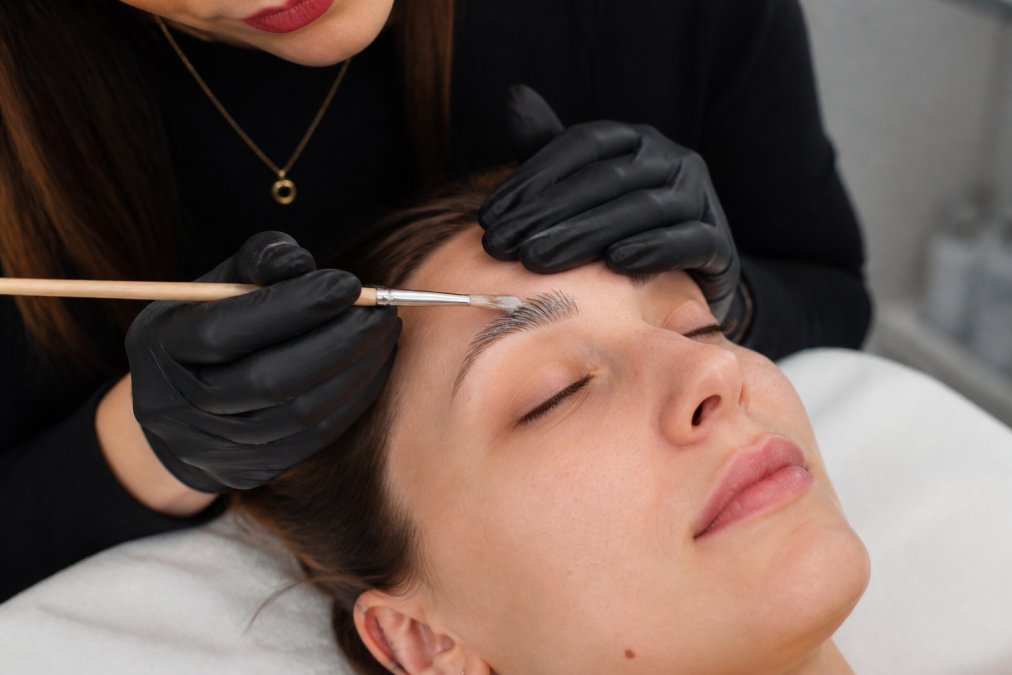 Professional applying lifting solution to eyebrows with precision brush, brow hairs visibly lifted and separated, clinical lighting showing chemical application technique
