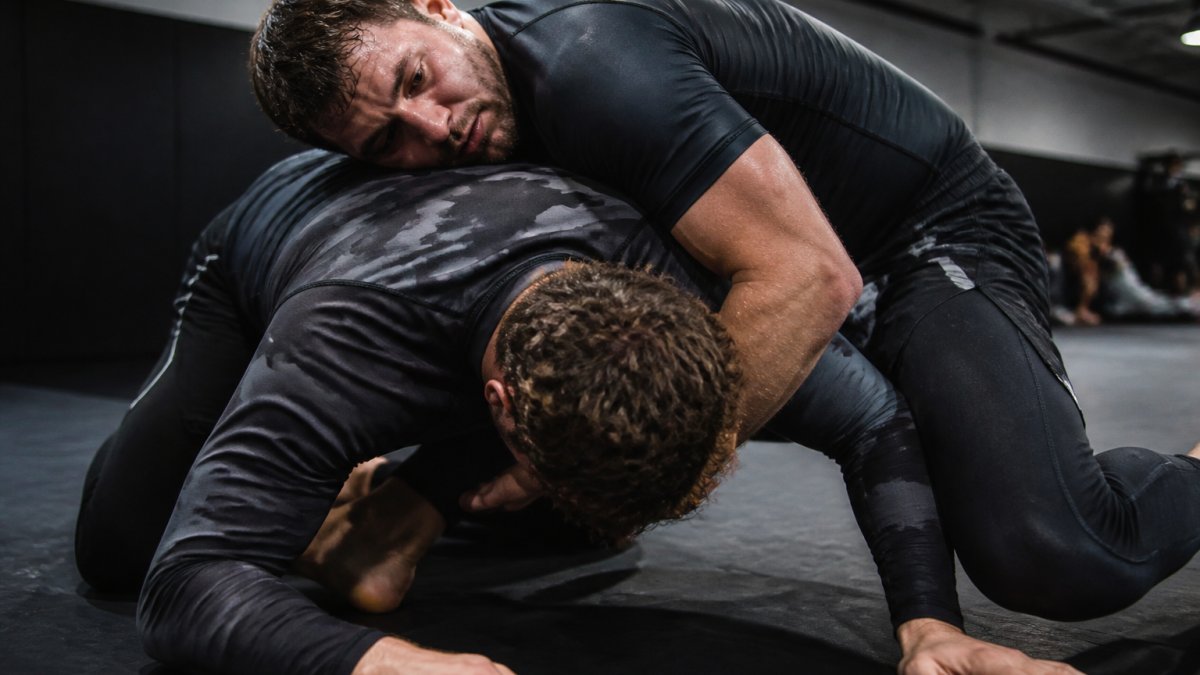 Grappler executing takedown on mat during intense training session, sweat visible, focused expression, gym lighting