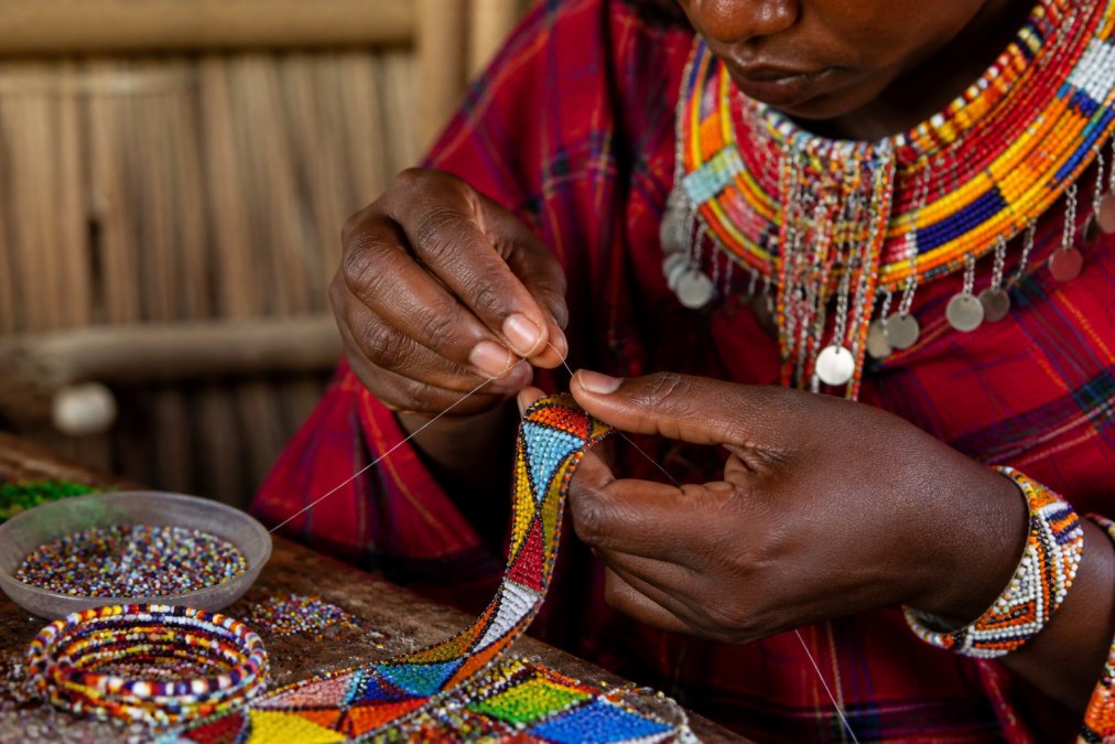 Close-up of artisan hands working with colorful beads and thread, weaving intricate patterns into traditional East African jewelry with focused concentration and natural workshop lighting