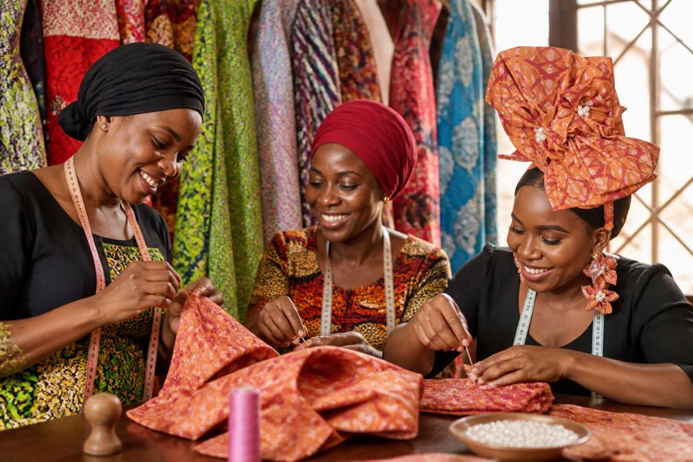 Female artisans working together at a textile workshop, hands engaged in detailed handcrafting with vibrant fabrics, warm natural lighting highlighting the intricate beadwork and stitching