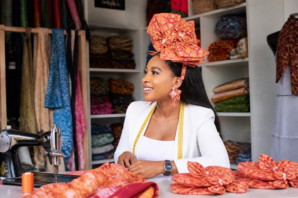 A confident woman artisan in professional attire smiling in her workspace surrounded by colorful handcrafted textiles and finished pieces, representing economic empowerment and pride in her craft