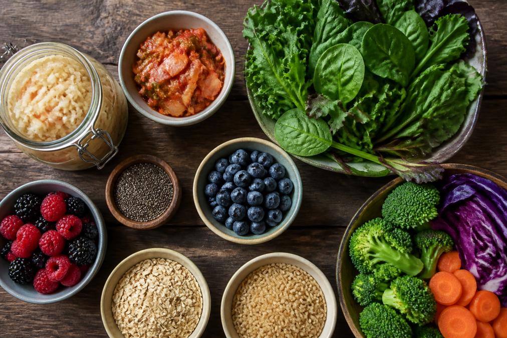 Overhead shot of colorful fermented foods and fiber-rich vegetables on a wooden table: sauerkraut in a glass jar, kimchi, fresh leafy greens, berries, and whole grains arranged naturally