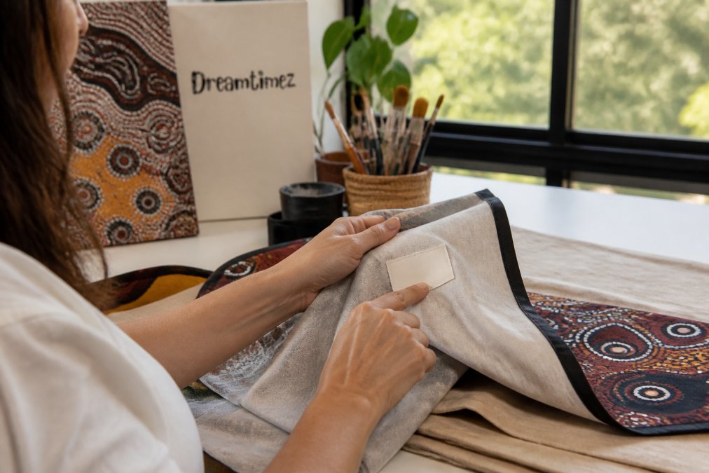 Person's hands examining a textile artwork with detailed Indigenous patterns, pointing to an artist attribution label visible on the back, natural daylight from window