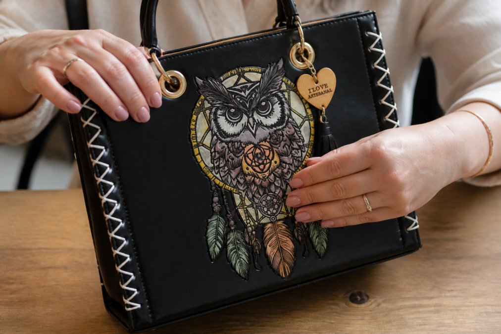 Close-up of hands examining a leather handbag with visible hand-painted floral details, showing texture variations and artisan brushwork on natural tan leather