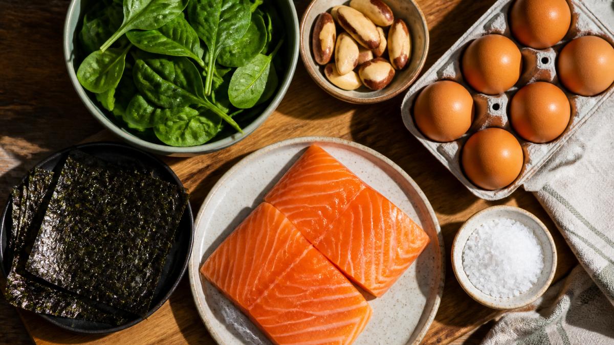 Overhead flat lay of thyroid-supporting foods including fresh salmon fillets, free-range eggs, seaweed sheets, Brazil nuts, leafy spinach, and iodised salt on a wooden board with natural morning sunlight
