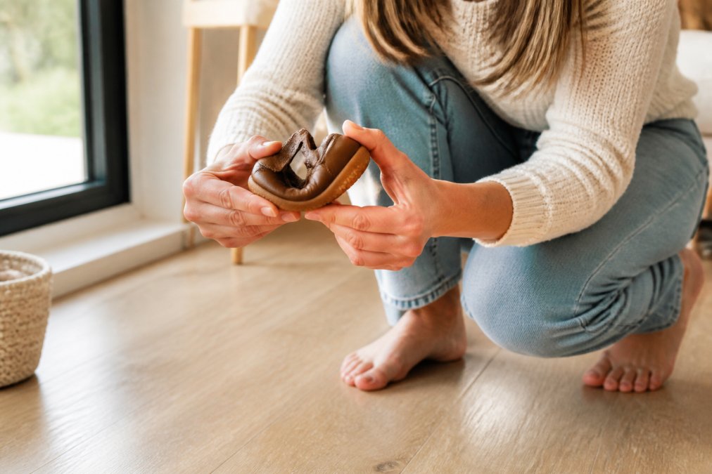 A parent crouching on a light wooden floor, gently flexing the sole of a small leather toddler shoe between both hands, soft daylight from a nearby window