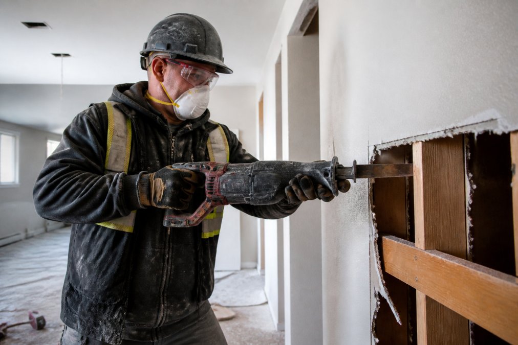 A contractor in full PPE, safety glasses, dust mask, and gloves, using a reciprocating saw to carefully remove a section of interior drywall while adjacent finishes remain intact