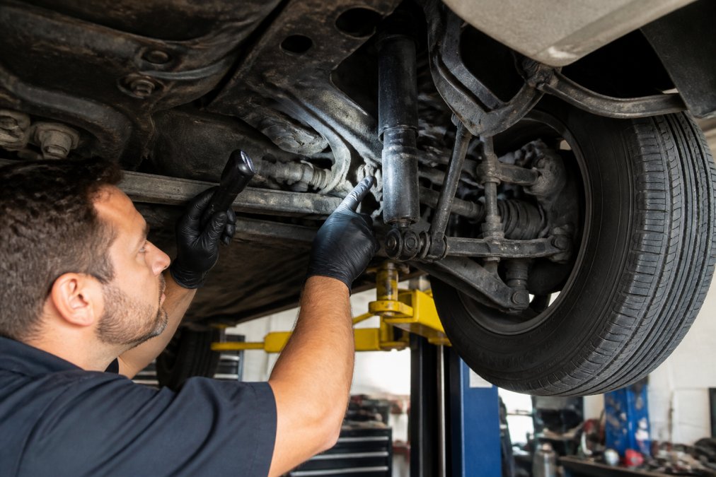 A certified technician inspecting a car's suspension system on a lift in a clean, well-lit auto repair shop, pointing at a worn shock absorber with a flashlight