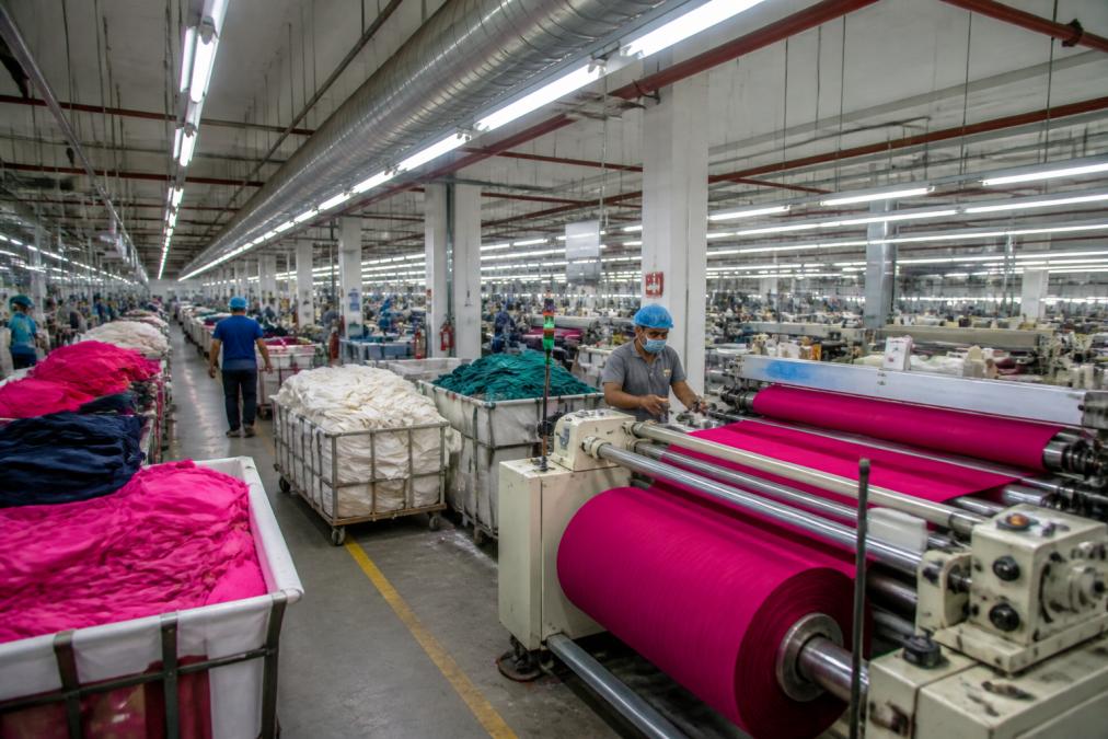 Large textile manufacturing facility with industrial machinery, rolls of colorful fabric on production lines, workers in safety gear managing garment production under bright overhead lighting