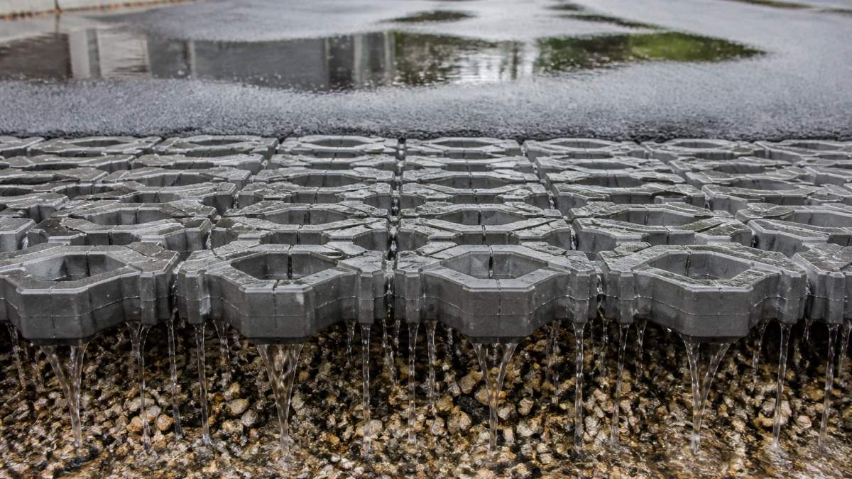 Close-up of water flowing through transparent permeable paver grid system into gravel base, with traditional asphalt showing standing water puddles in the background