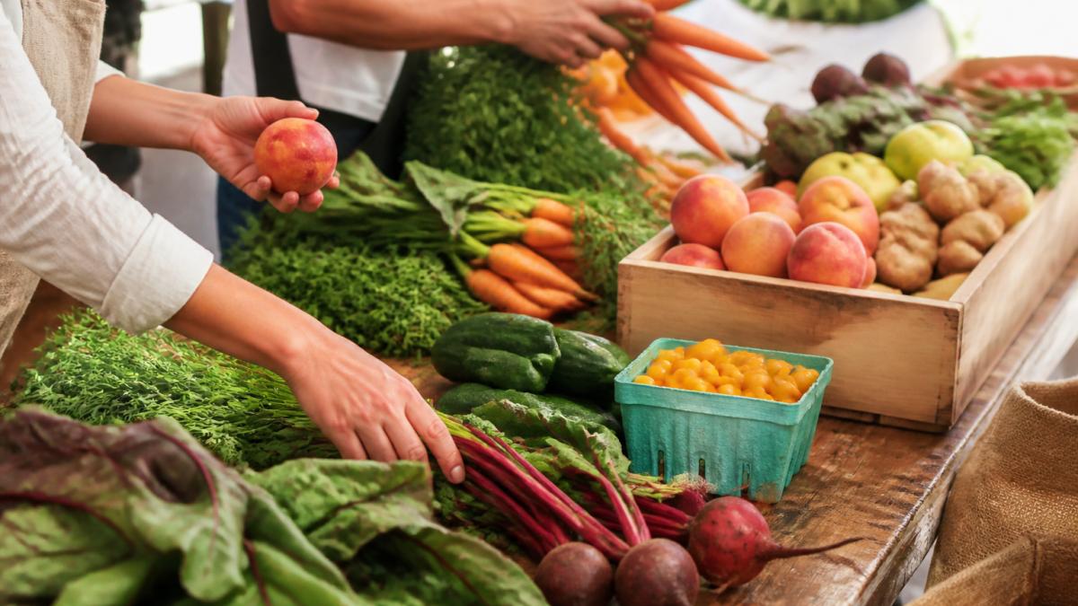 Colorful fresh fruits and vegetables from farmers market being sorted and arranged on wooden work surface in bright natural light, with hands selecting ripe produce for processing into beverages