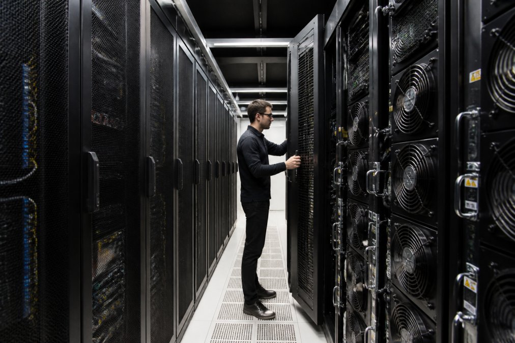 A technician in a data center aisle inspecting a modern in-row cooling unit installed between server racks, with visible airflow vents and clean cable management