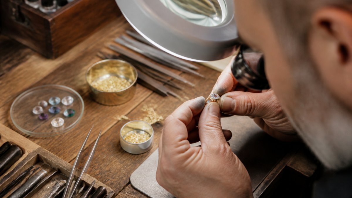 Jeweler's hands carefully examining recycled gold and gemstones under magnification lamp on wooden workbench with precision tools