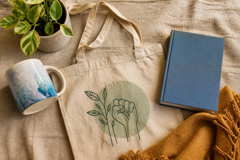 A flat lay of activist-themed gifts arranged on a natural linen surface: an artisan ceramic mug with a watercolor design, an organic cotton tote bag, a hardcover book, and a small potted plant, warm natural lighting