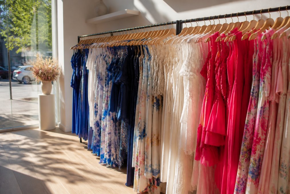 A neatly arranged rack of colourful designer dresses in a sunlit boutique, natural light streaming through a large window highlighting the fabric textures and varied silhouettes, the space clean and carefully curated