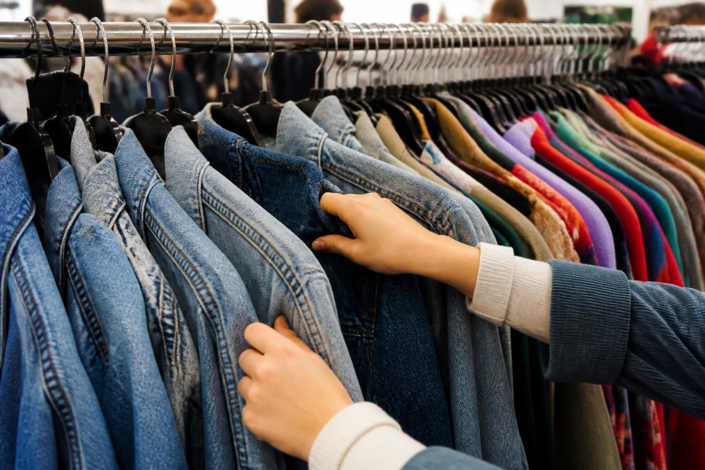 Close-up of hands flipping through vintage denim jackets and colorful clothing on racks at a thrift store, showing fabric textures and color variety with natural lighting
