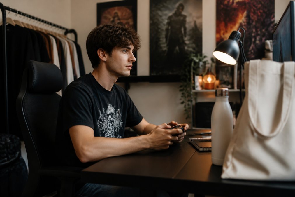 A young adult wearing a graphic gaming t-shirt sitting at a desk with a computer and gaming controller, surrounded by a reusable water bottle and canvas tote bag, in a cozy room with gaming posters and warm desk lighting