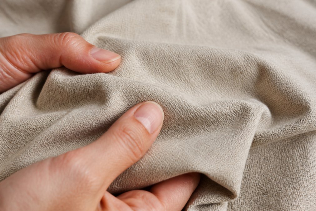 Close-up of organic cotton fabric texture showing natural fibres and heavier-knit weave in neutral colour, hands gently touching the material to show durability and premium feel