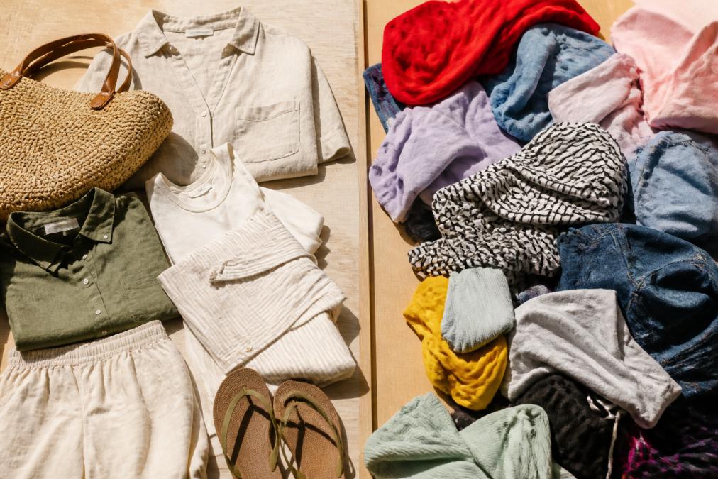 A neatly arranged flat lay of quality coastal-style clothing items in neutral tones, linen shirts, soft cotton tops, and a woven tote, on a light wooden surface in warm afternoon sunlight