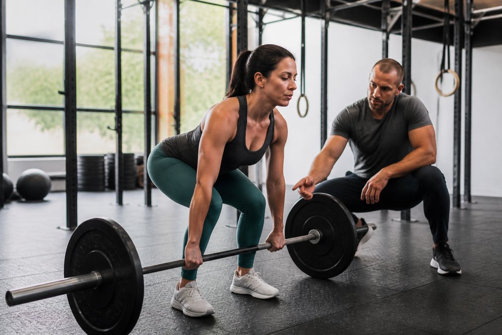 A woman in her 30s performing a barbell deadlift in a well-lit gym, focused expression, wearing athletic leggings and a fitted top, with a coach observing nearby and offering form cues