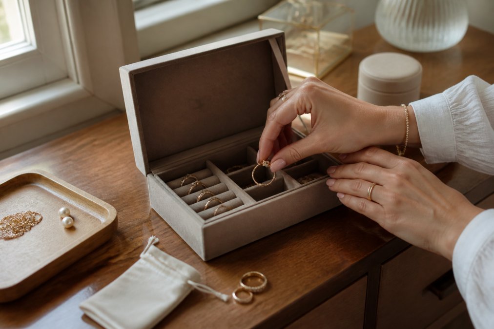 Close-up of a woman's hands placing a gold ring into a velvet-lined jewelry box compartment, soft natural window light on a wooden dresser