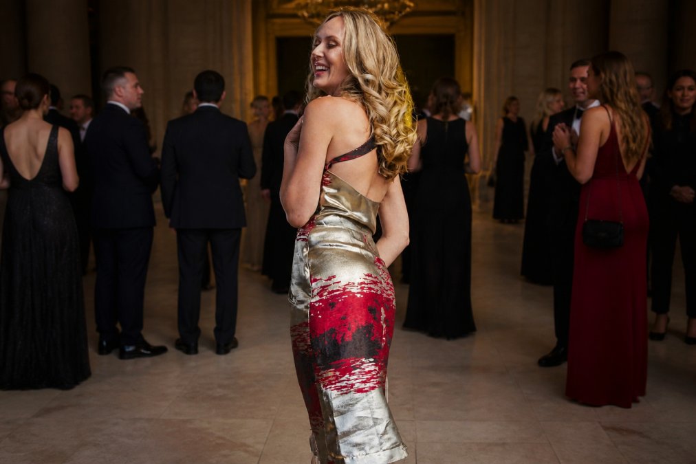 Woman wearing a burgundy gold metallic jacquard halter dress at an elegant evening event, showing the luminous quality of the fabric under soft lighting