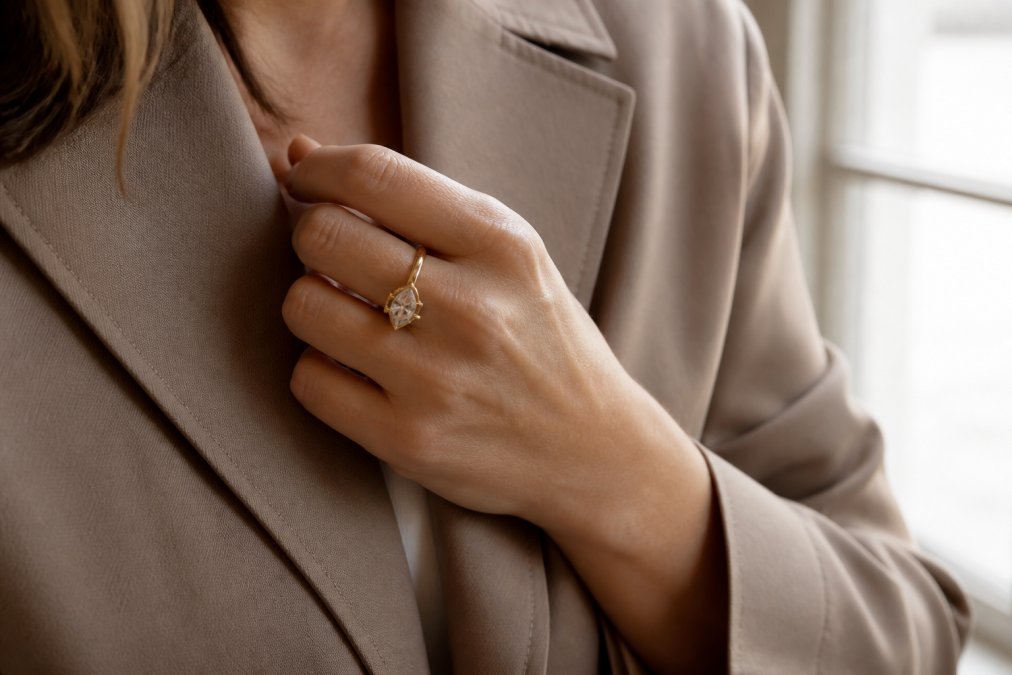 Close-up of a woman's hand wearing a bold 18k gold ring with an antique marquise diamond, styled with a neutral-toned blazer, photographed in soft natural window light that catches the metal's warm shine